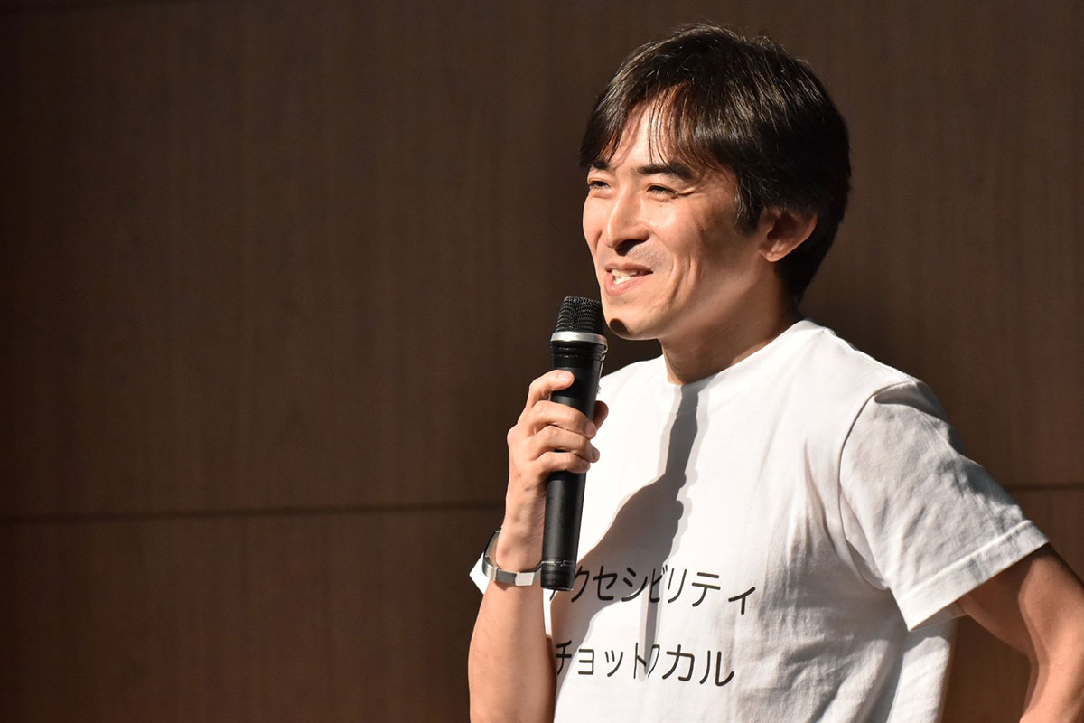 写真:笑顔でお話中の木達さん。Tシャツは白地に黒文字