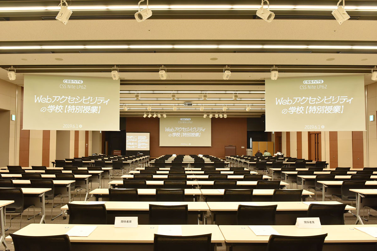 写真:整然と机と椅子が並んだ会場。前方中央と後方左右にはスライドを写すスクリーンがある。