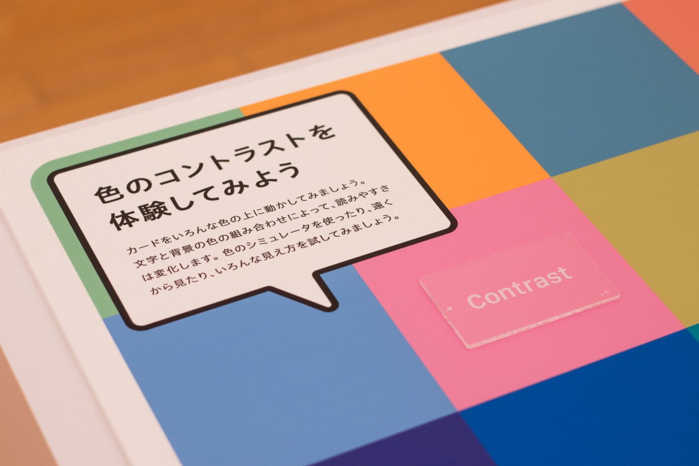 写真:「色のコントラストを体験してみよう」のコーナー。カラフルなパネルの上にContrastという白い文字が書かれたアクリルカードが載っています。
説明文:カードをいろんな色の上に動かしてみましょう。文字と背景の色の組み合わせによって、読みやすさは変化します。色のシミュレータを使ったり、遠くから見たり、いろんな見え方を試してみましょう。
