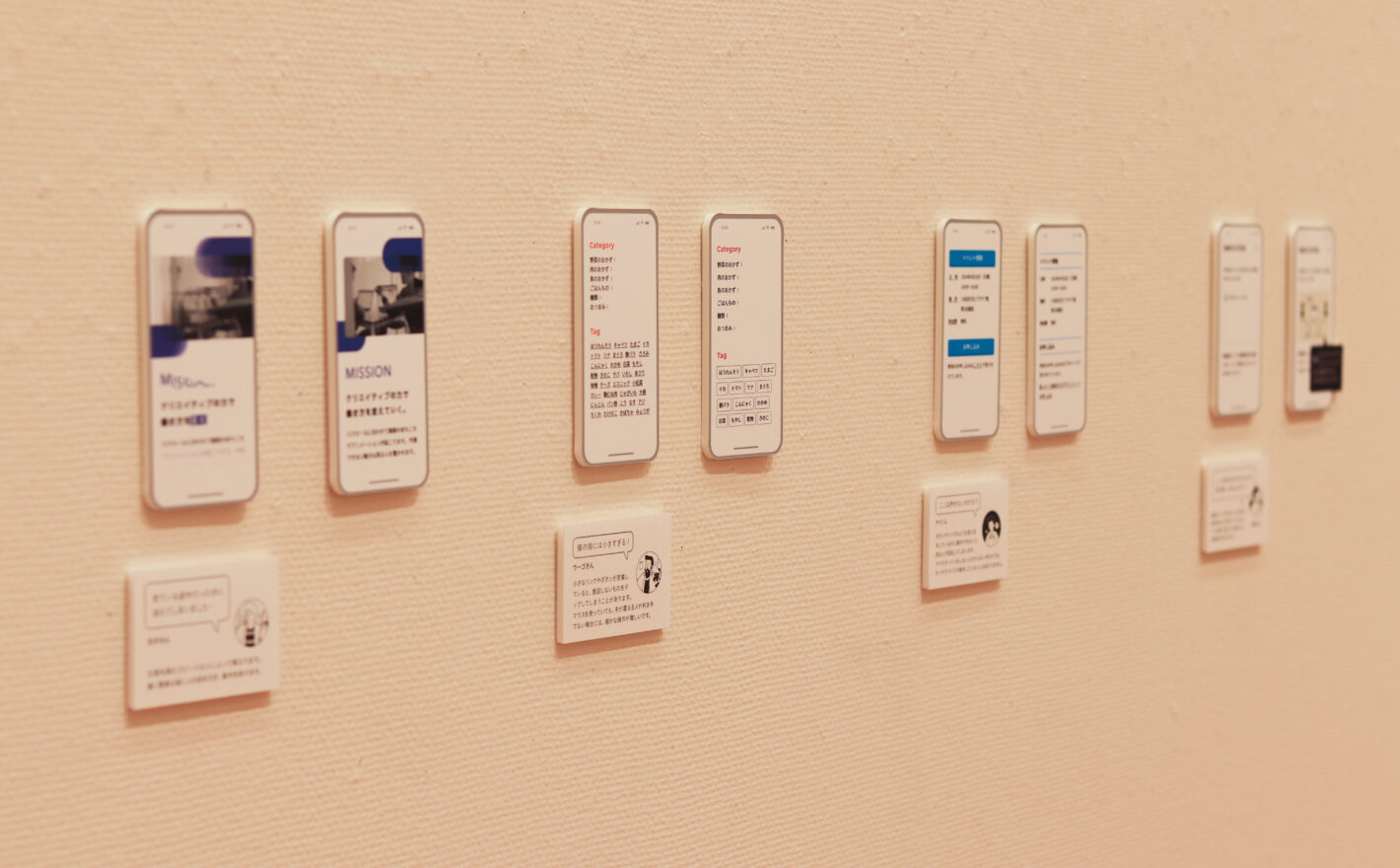写真:スマホサイズのモックアップが4組、壁に展示してあります。それぞれ下に登場人物のセリフ入りキャプションがついています。