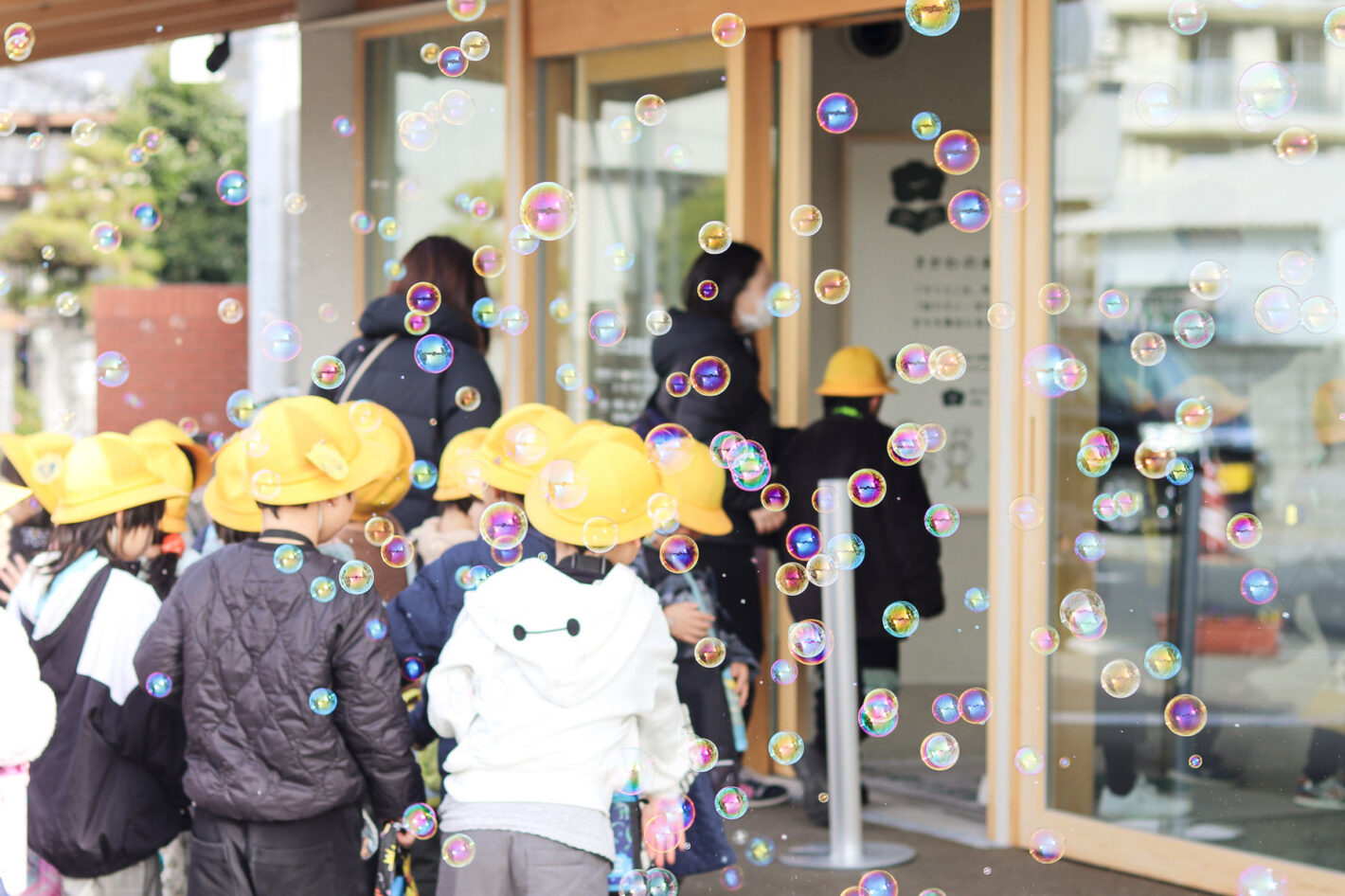 写真:黄色い帽子をかぶったこどもたちが、たくさんのシャボン玉のなかをくぐって図書館へ入っていくところ。